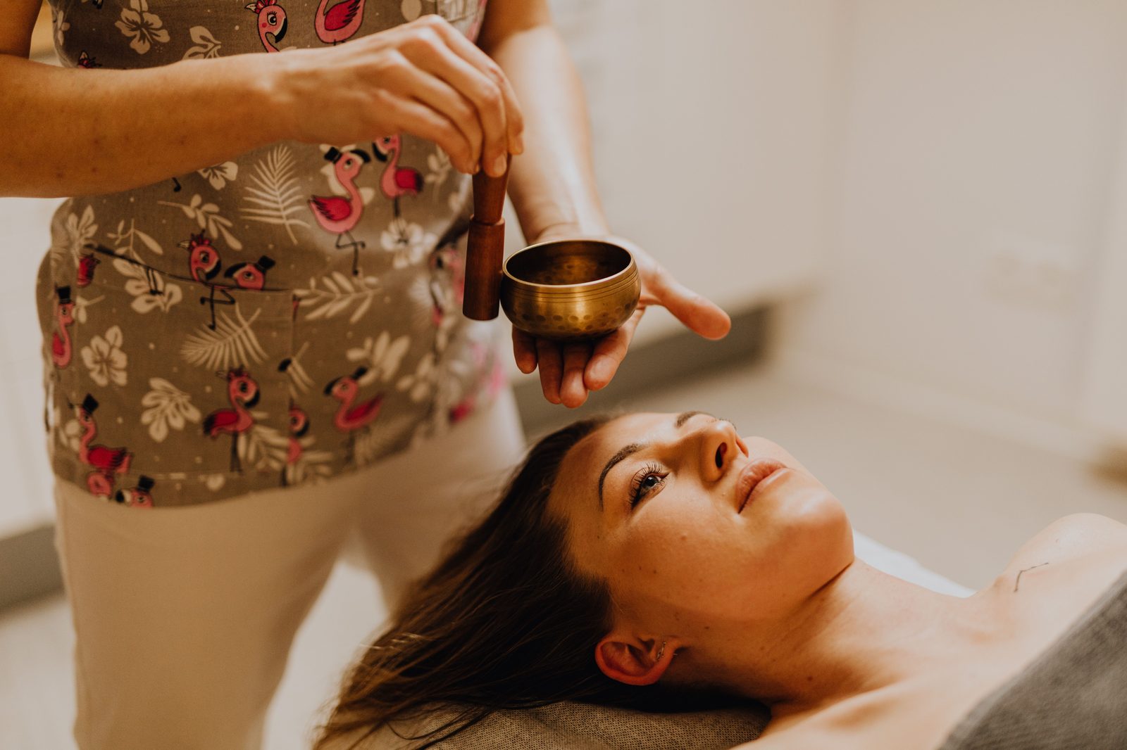 Client relaxing on a spa table while a therapist holds a singing bowl during a Sound Bath Facial